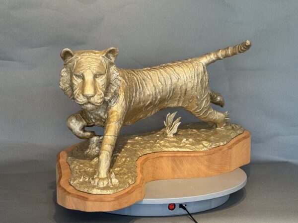Naranji is a golden tiger sculpture with one paw raised, mounted on a wooden base shaped to fit the figure and displayed against a plain gray background.