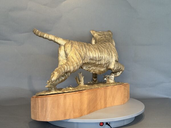 The Naranji is a golden tiger sculpture on a wooden base, shown from behind in mid-stride. Its detailed muscle and fur textures stand out against a plain gray background.