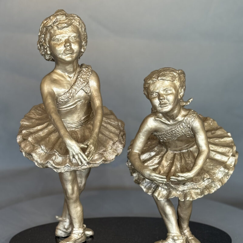 Showtime I and II: bronze sculptures of two young ballerinas in tutus and ballet slippers, gracefully posed on a black oval base against a neutral background.