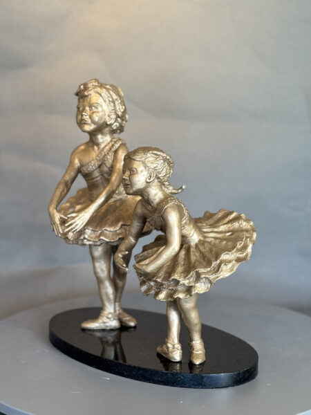 The Showtime I and II bronze sculpture depicts two young ballerinas in tutus on a black oval base—one stands upright with arms lowered, while the other bends forward, both gracefully posed in dance positions.