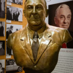 A bronze bust of Sidney S. Kellam in a suit and tie is displayed in a museum, with black-and-white photos and a large poster featuring his name and portrait behind it.