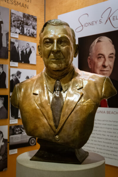 A bronze bust of Sidney S. Kellam in a suit and tie is displayed in a museum, with black-and-white photos and a large poster featuring his name and portrait behind it.