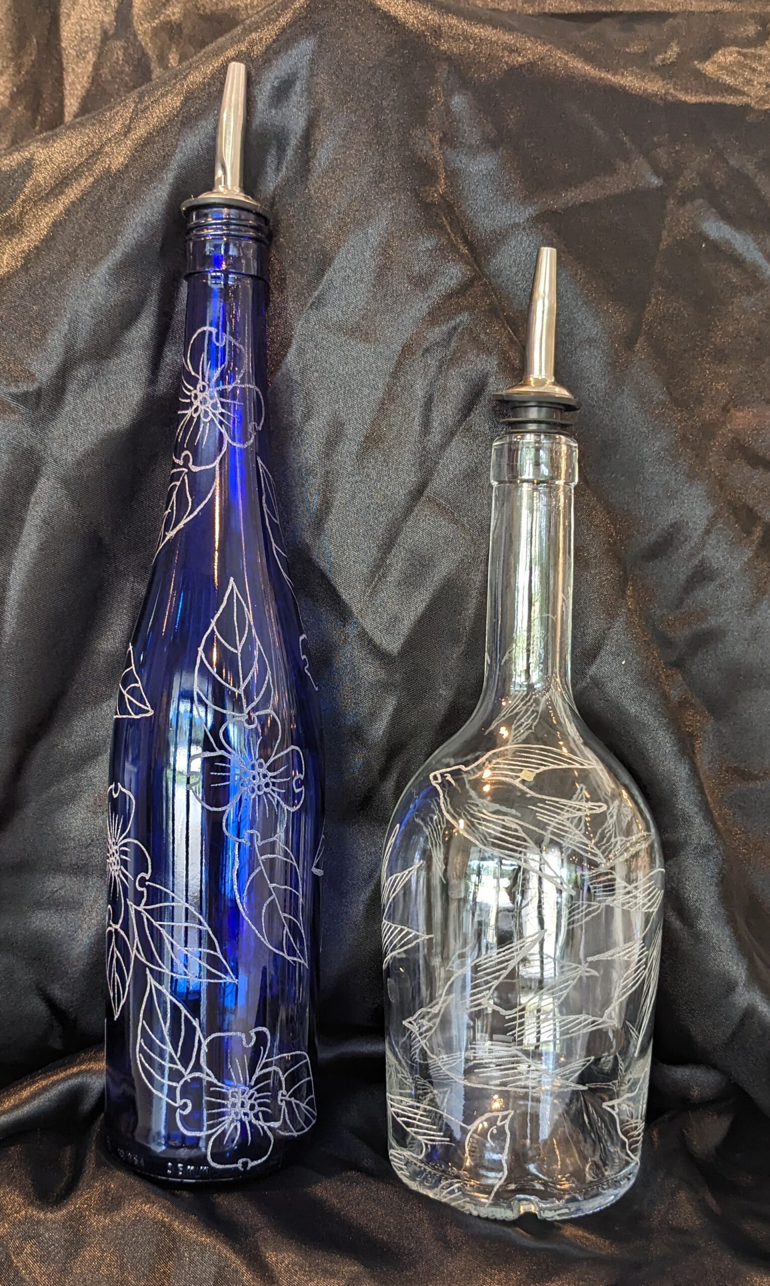 Two Olive Oil/Soap Bottles with metal pour spouts are shown against a black fabric background. One is blue with white floral designs; the other is clear, featuring etched leaves and birds.