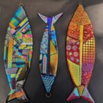The Dichroic Fish are three colorful, fish-shaped mosaics crafted from iridescent and patterned glass pieces. Each features vibrant colors and geometric designs, showcased against a dark background.