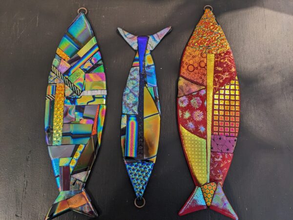 The Dichroic Fish are three colorful, fish-shaped mosaics crafted from iridescent and patterned glass pieces. Each features vibrant colors and geometric designs, showcased against a dark background.