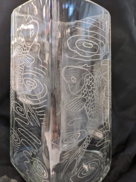 The Large Koi Vase is a clear glass vase featuring etched koi fish and swirling water designs, beautifully displayed against a dark background.