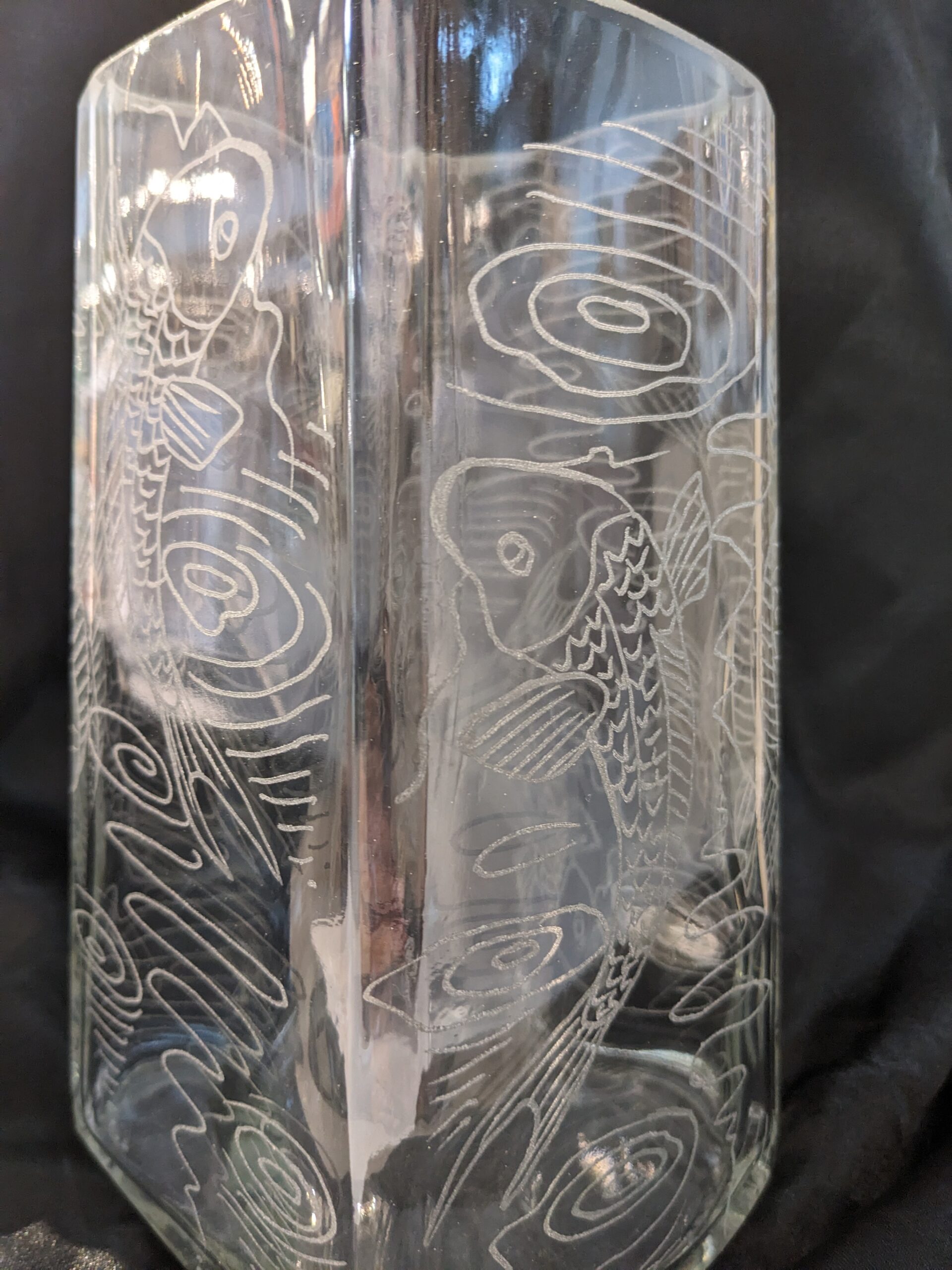 The Large Koi Vase is a clear glass vase featuring etched koi fish and swirling water designs, beautifully displayed against a dark background.