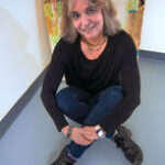 A woman with shoulder-length blonde hair, wearing a black sweater, blue jeans, and black shoes, sits cross-legged on a gray floor and smiles at the camera. A colorful abstract painting is in the background.